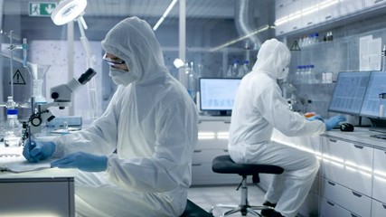 In a Secure High Level Laboratory Scientists in a Coverall Conducting a Research. Biologist Adjusts Samples in a  Petri Dish with Pincers and then Examines Them Under Microscope. - Powered by Adobe