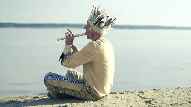 An Indian in a national costume plays a flute on the river bank 4k