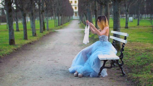Attractive girl in silver and blue dress sits on bench in parkway adjusting dress, puts on white tank top and gets ready to pose during photo shoot.