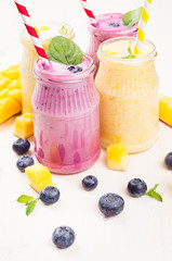 Freshly blended yellow and violet  fruit smoothie in glass jars with straw, mint leaves, mango slices, blueberry, close up. Soft white wooden board background.