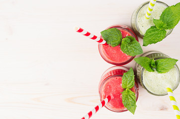 Freshly blended green and red fruit smoothie close up in glass jars with straw, mint leafs, top view. White wooden board background, copy space.