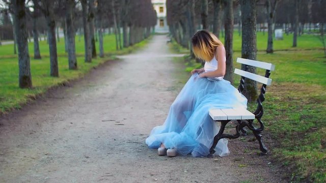 Attractive girl in silver and blue dress sits on bench in parkway changing clothes and getting ready to pose during photo shoot.