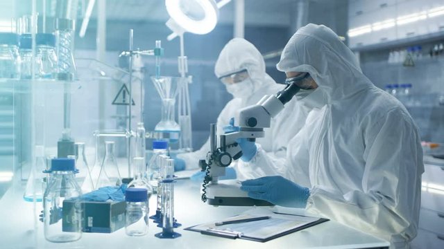  In a Secure High Level Laboratory Scientists in a Coverall Conducting a Research. Chemist Adjusts Samples in a  Petri Dish with Pincers and then Examines Them Under Microscope.