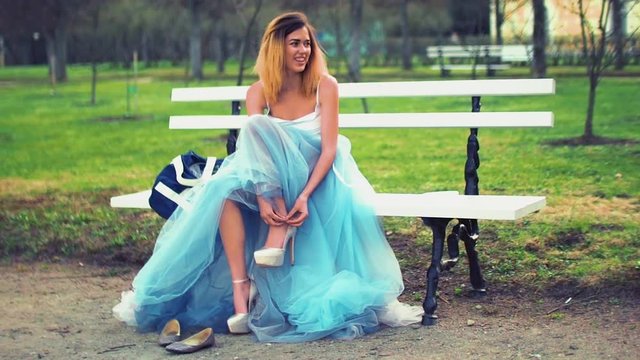 Attractive girl in silver and blue dress sits on bench in parkway putting on high heeled shoes and gets ready to pose during photo shoot. Front view.