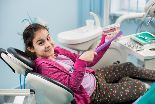Smiling girl in dentist chair showing proper tooth-brushing using dental jaw model and big toothbrush in dental office. Dentistry, early prevention, oral hygiene concept