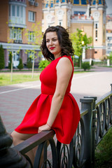 Fototapeta na wymiar Beautiful and attractive european young woman in red dress and black jacket walk in city near park and fountain. Have a pretty makeup and red lips