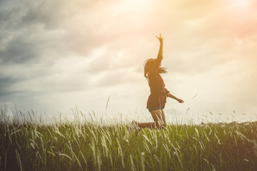 Woman jumping and raising up her hand and two finger over grass field on summer evening