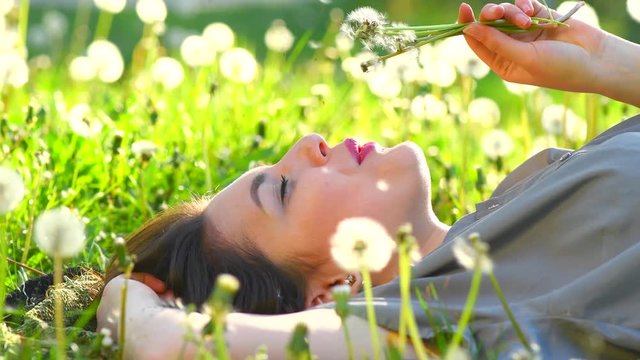 Beautiful young woman lying down on the field in green grass, blowing dandelions and smiling