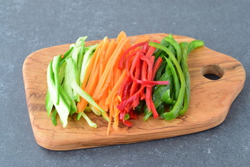 Fresh cucumber, carrot, red and green sweet paprika sliced in stripes on an olive wood cutting board on a grey abstract background. Step by step cooking
