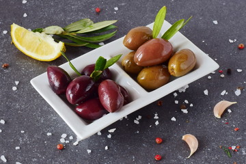 Black and green olives in a white plate on a grey abstract background with lemon, garlic, spices. Mediterranean food. Healthy eating concept