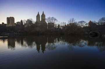 Naklejka na ściany i meble Scenic sunset view of the New York City skyline with bare winter trees reflecting with buildings of the Upper West Side in the Central Park lake