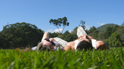 Young couple lying on green grass in park and relaxing. Man and woman sitting on meadow at nature and kissing. Girl and boy looking at the landscape and enjoying vacation. Low angle of view Rear Back