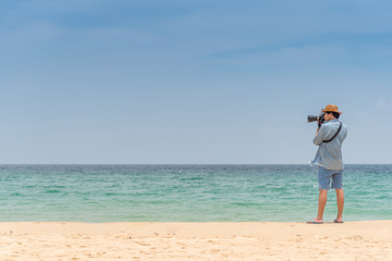 young Asian man photographer with jean shirt and hat take photo of tropical island beach and turquoise sea, seascape background for summer holiday and vacation travel concepts