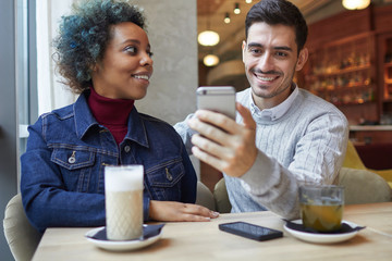 Closeup of young mixed couple enjoying their time together in coffee house sitting together with drinks in front of them. Guy is holding smartphone to take photo, girl is looking at him with smile.