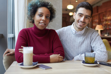 Indoor shot of African American girl and Caucasian guy sitting together at one cafe table in...