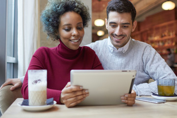 Indoor shot of happy mixed couple enjoying their date in small coffee house sharing media content with each other, looking together with interest and happy smiles at display of tablet computer.