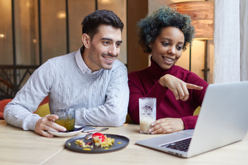 Portrait of happy lovers meeting indoors with laptop open on table in cafe. Dark-skinned girl is pointing to display with joyful smile, man is looking attentively at screen involved in what she shows.