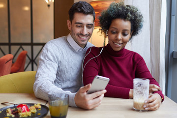 Loving mixed couple staying in cafe, European male hugging his ethnic girlfriend, they are both listening to audio tracks from his smartphone sharing earphones, smiling, looking happy and pleased.