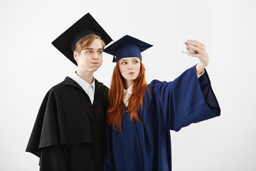 Two cheerful graduates of university fooling making selfie over white background.