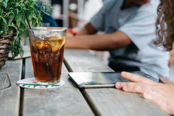 Refreshing glass of cola on wooden table in a bar while young is connected with tablet