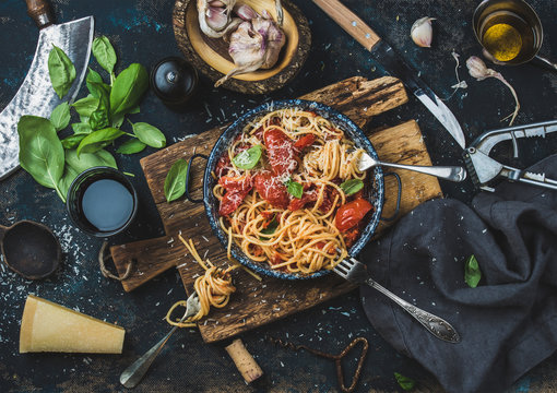 Italian style pasta dinner. Spaghetti with tomato and basil in plate on wooden board and ingredients for cooking pasta over dark blue plywood background, top view