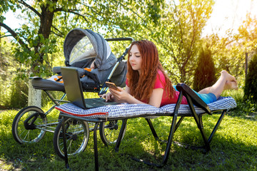 Young woman with her newborn baby lying on the soft armchair with laptop and looking at the phone. Summer day. Mother with child outdoors. Motherhood.