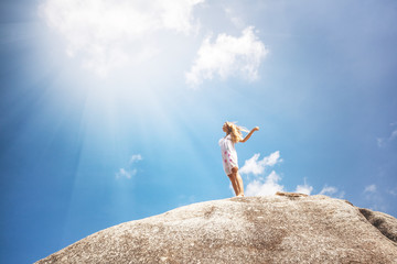 Young woman in white dress is standing on a high stone against the blue sky and sun rays