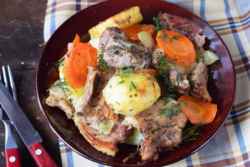 Oven cooked pork with potato, carrots and onion in olive oil and spices in a brown ceramic plate on a wooden background. Home cooking