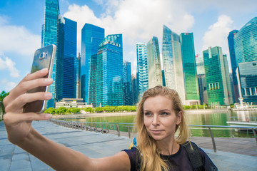 Fototapeta na wymiar Girl holding smartphone for self-portrait photo with view of modern skyscrapers during summer travel vacation