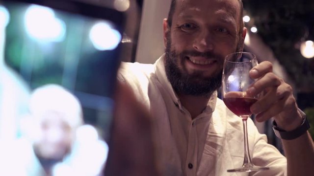 Young man taking selfie photo with cellphone and drinking red wine in cafe at night 
