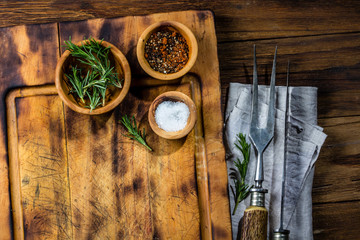 Cutting wooden board, cutlery set, rustic linen napkin, and spices on wooden background