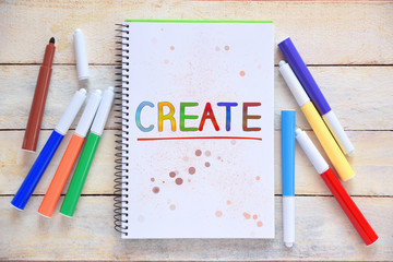 Some markers on a white wooden desktop and a notebook with the word CREATE colorful hand written on it. Empty copy space for Editor's text.