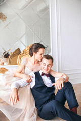 Bride in beautiful dress and groom in black suit sitting on sofa indoors in white studio interior like at home. Trendy wedding style .