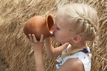 Girl drinks cow's milk from a clay pot against the backdrop of a haystack on a farm.