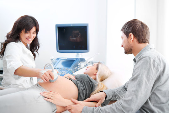 Cheerful female doctor smiling during ultrasound scanning procedure for her pregnant patient and her loving husband couples medicine hospital clinic diagnosis technology family childbirth profession.