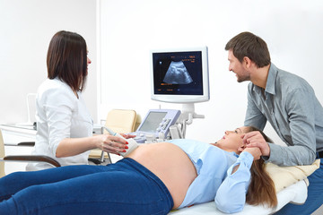 Obraz premium Pregnant young woman holding hand of her loving husband during medical examination at the clinic. Female doctor performing ultrasound scanning on her pregnant patient pregnancy fertility expecting.