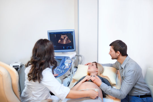 Happy loving pregnant couple visiting doctor together. Gynecologist performing ultrasound scanning for her pregnant patient. Mature man holding hand of his pregnant wife during medical examination.
