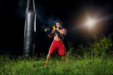 Powerful strong young muscular shirtless boxer with burning gloves training outdoors on a punching...