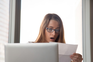 Surprised young businesswoman reading letter at workplace. Puzzled office worker looking on document in hand. Female entrepreneur receiving unexpected official notice or personal invitation