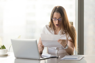 Surprised young businesswoman reading letter at the desk in front of laptop. Woman feels shocked after receiving unexpected news in written message. Female entrepreneur holds notice about loan debt