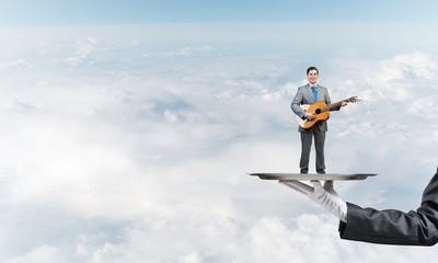 Businessman on metal tray playing acoustic guitar against blue sky background