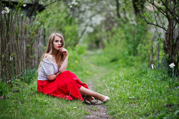 Portrait of sitting beautiful girl with red lips at spring blossom garden on green grass, wear on red dress and white blouse.