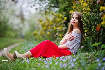 Naklejka na ściany i meble Portrait of sitiing beautiful girl with red lips at spring blossom garden on grass with flowers, wear on red dress and white blouse.