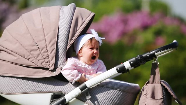 Crying baby-girl sitting in the baby carriage in the park at sunlight day.