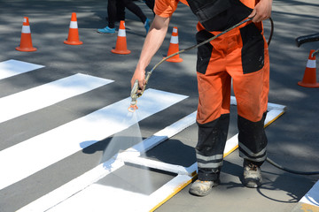 Worker is painting a pedestrian crosswalk. Technical road man worker painting and remarking pedestrian crossing lines on asphalt surface using paint sprayer gun.