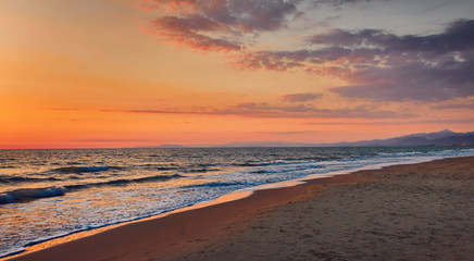 Panorama of a deserted beach at sunset. Turquoise foam waves on a sandy beach, golden sky with clouds from the setting sun, foggy blue silhouette of mountains in the background