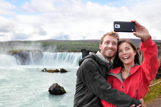 Selfie couple taking smartphone picture of Godafoss waterfall outdoors on Iceland. Couple visiting famous tourist attractions and landmarks in Icelandic nature landscape. Mixed race couple having fun.