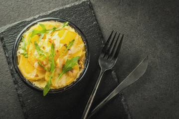 Casserole. French cuisine. Homemade potato gratin in a bowl for baking. On a black stone background. With leaves of fresh arugula for decoration. Top view copy space