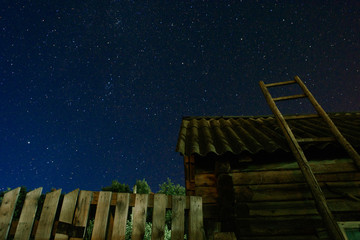 Village. Old wooden ladder leaning against the barn with a slate roof in the night star sky. The staircase leads to the sky.