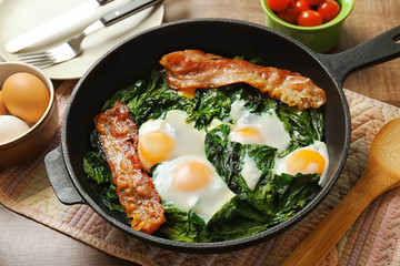 Frying pan with tasty eggs, spinach and bacon on wooden table